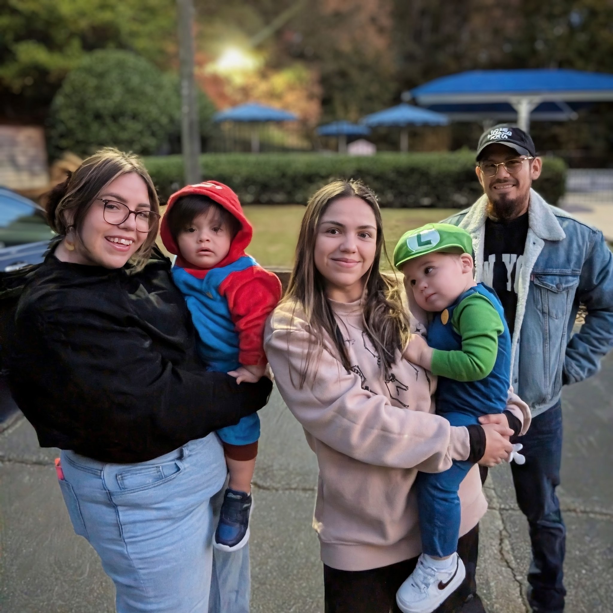 First Halloween trick-or-treating with my grandchildren — the family group at dusk, both toddlers in Mario and Luigi costumes, Fri · Oct 31, 2025